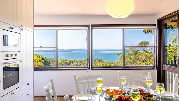 A dining area with a table set for four, overlooking a scenic view of the sea and hills. - Casa Carinho, 24 Wallawa Rd - spectacular water views, pet friendly, air con and Wi-Fi (Nelson Bay)