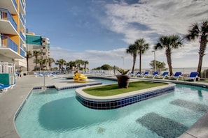 Una piscina con bordi piastrellati di blu, sedie a sdraio, palme e un edificio con balconi.