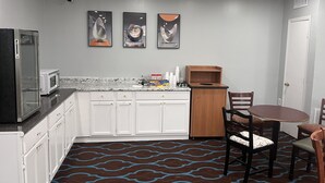 Cocina compacta con armarios blancos, microondas y refrigerador. Hay tres obras de arte enmarcadas en la pared, un pequeño mueble de madera y una mesa de comedor con sillas.
