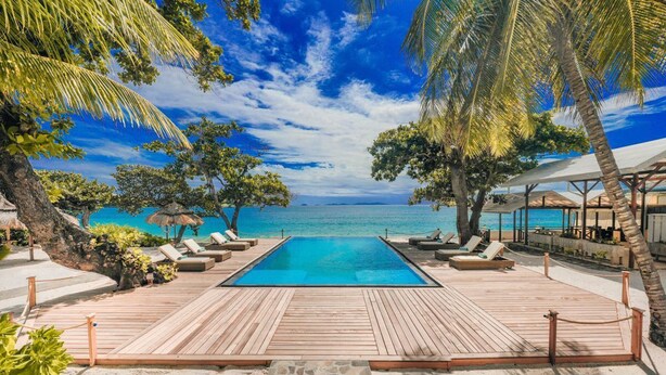 A wooden deck with a pool, lounge chairs, and umbrellas, overlooking a beach and ocean.
