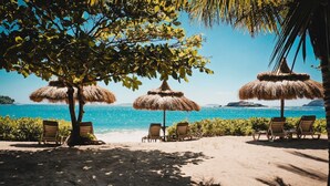A beach with thatched umbrellas and wooden chairs under trees, overlooking a clear blue sea.