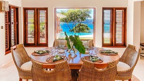 Un coin repas avec une table dressée pour quatre personnes, entouré de chaises en osier, et offrant une vue sur une piscine et des arbres à travers de grandes fenêtres.
