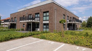 Un bâtiment moderne avec une façade en briques, de nombreux balcons et un parking dallé avec des places délimitées.