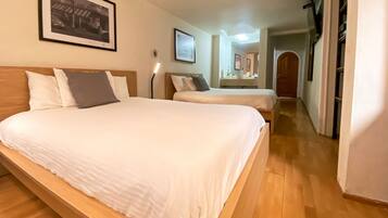 A hotel room with two beds, a wooden floor, a framed picture on the wall, and a hallway leading to a bathroom with a sink and mirror.
