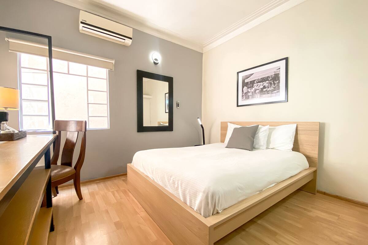 Een kamer met een houten bed, een bureau, een stoel, een airconditioner, een raam met jaloezieën en een ingelijst beeld aan de muur.