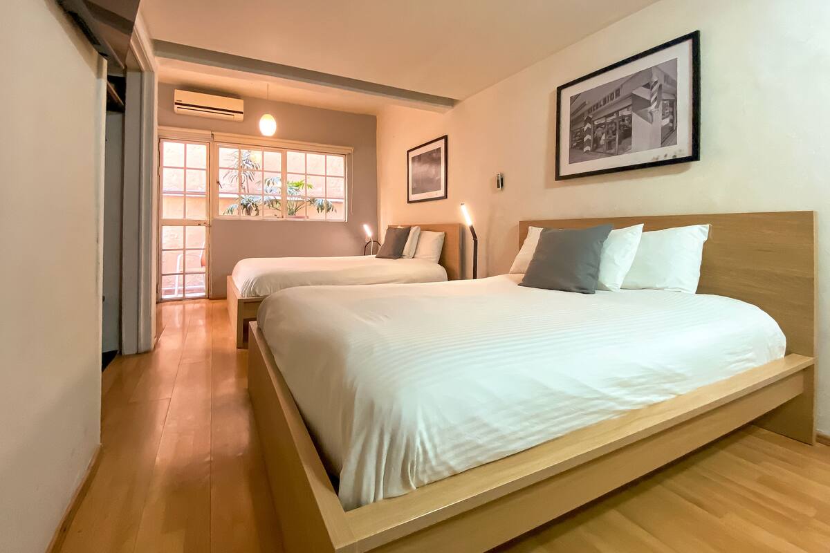 Een hotelkamer met twee bedden, een raam met planten, een lijst met afbeelding en een houten vloer.