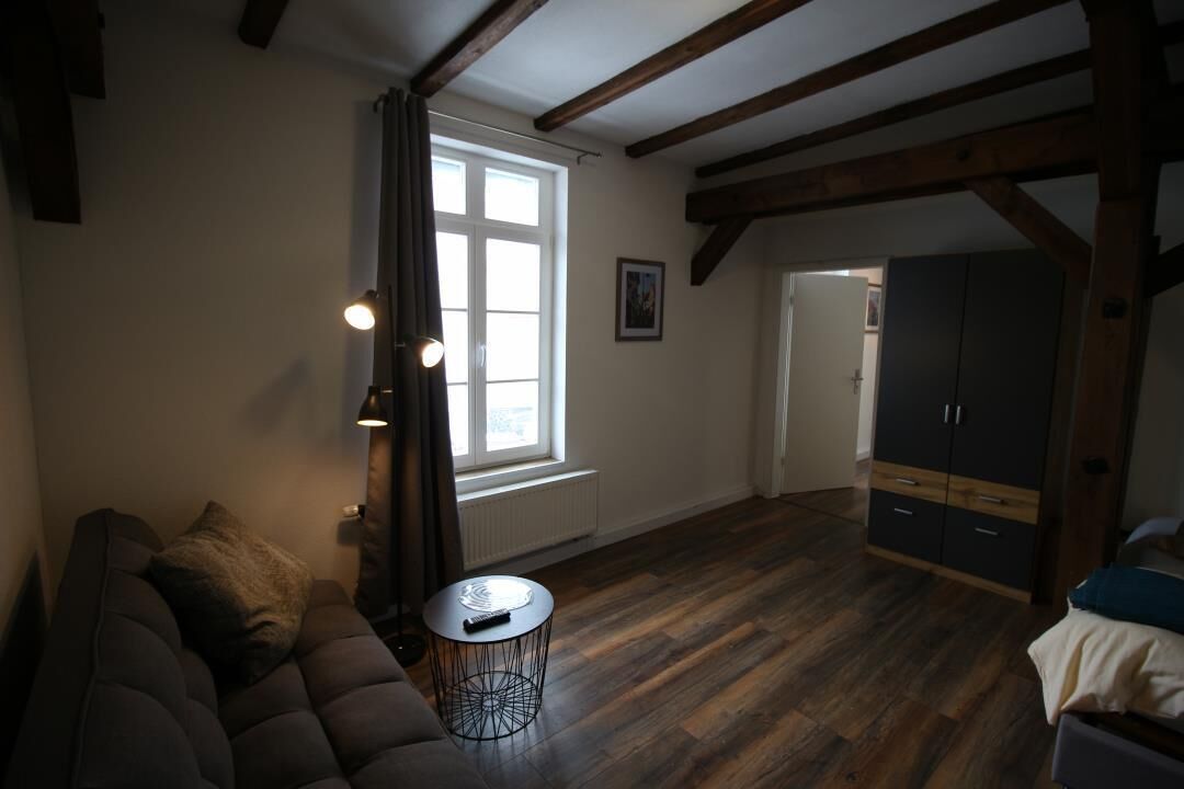 Una habitación con piso de madera oscura, un sofá, una mesita redonda, una ventana con cortinas y un armario.