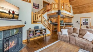 Un vaste espace de vie avec une cheminée, un escalier en bois, un canapé et une cuisine en arrière-plan.