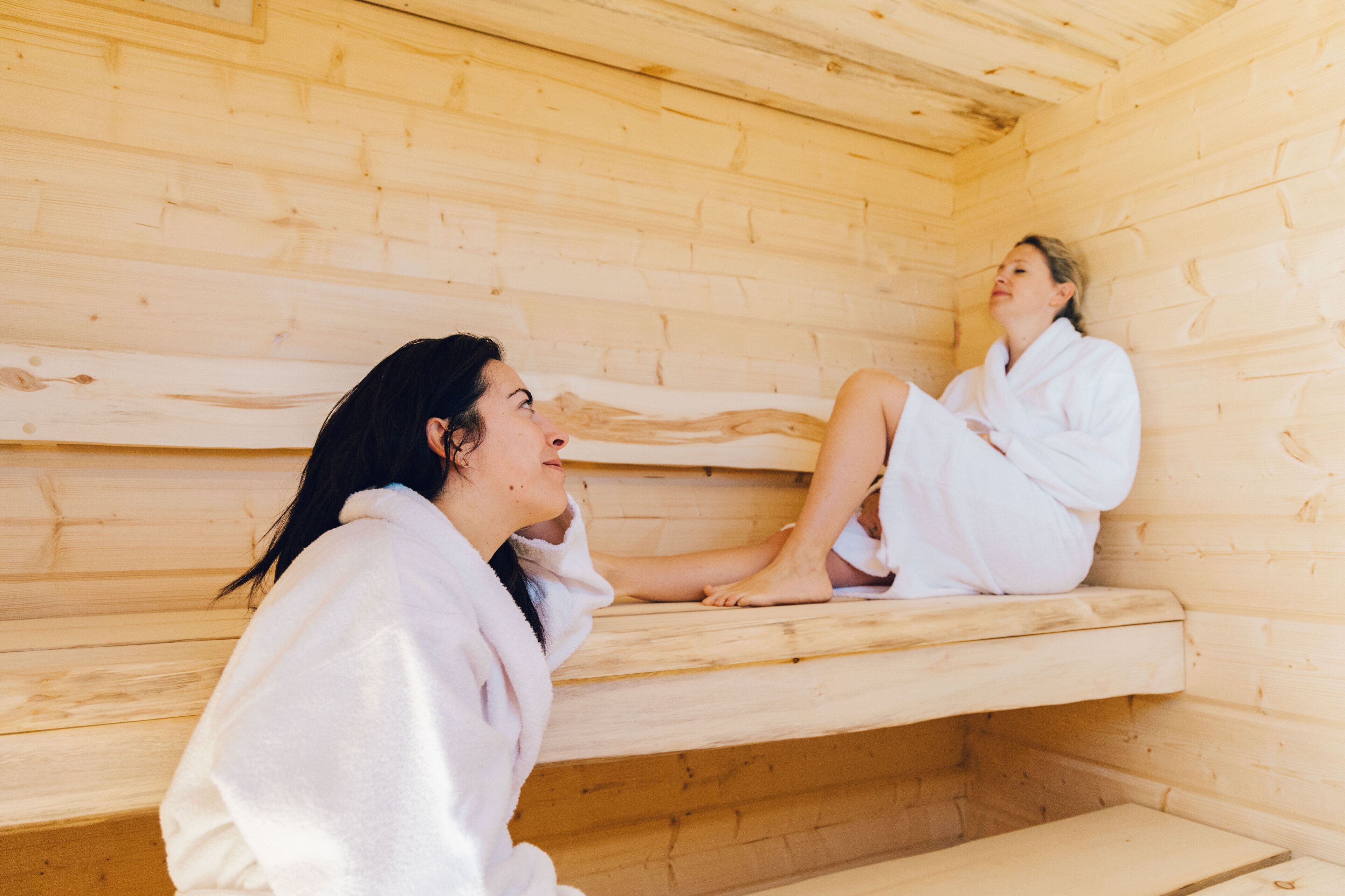 Two individuals in white robes seated in a wooden sauna cabin with benches.