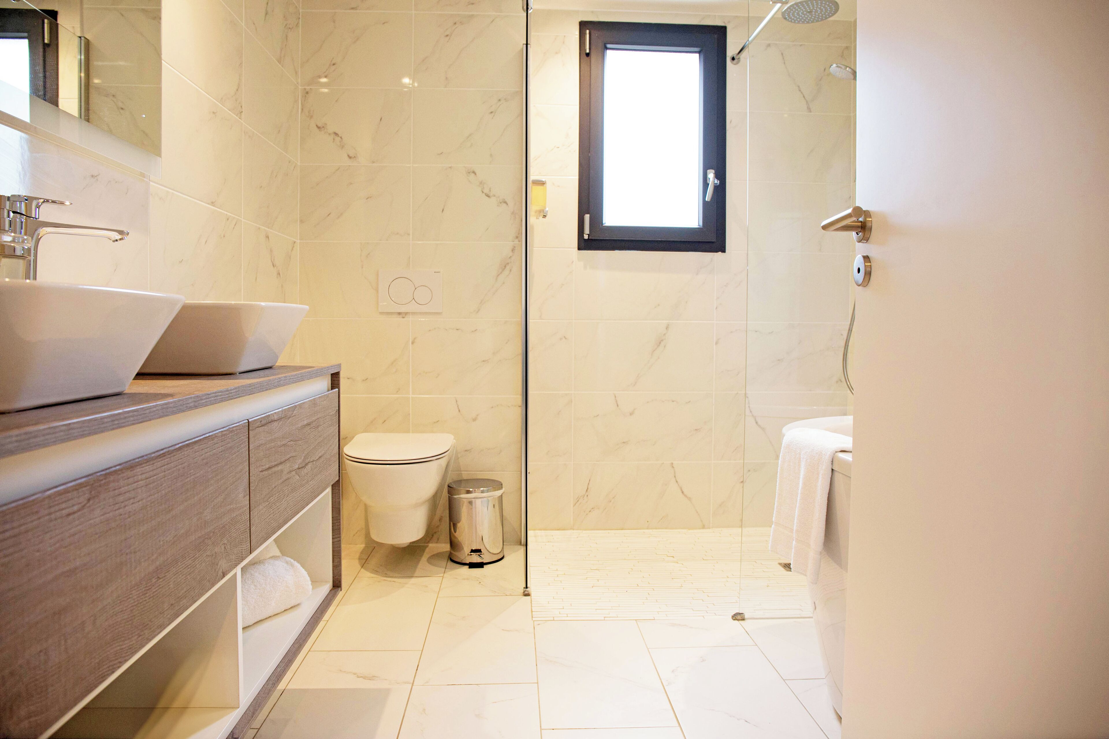Een moderne badkamer met een marmeren vloer, een houten wastafelmeubel met een grote wastafel, een toilet en een douchehoek.