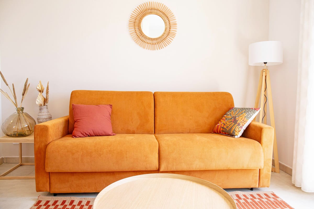 Una habitación con un sofá naranja, un espejo redondo, una lámpara y un jarrón con plantas secas.