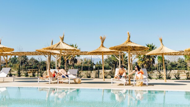 Un espace aménagé au bord de la piscine, avec des chaises longues et des parasols en chaume, entouré d’une clôture et de verdure.