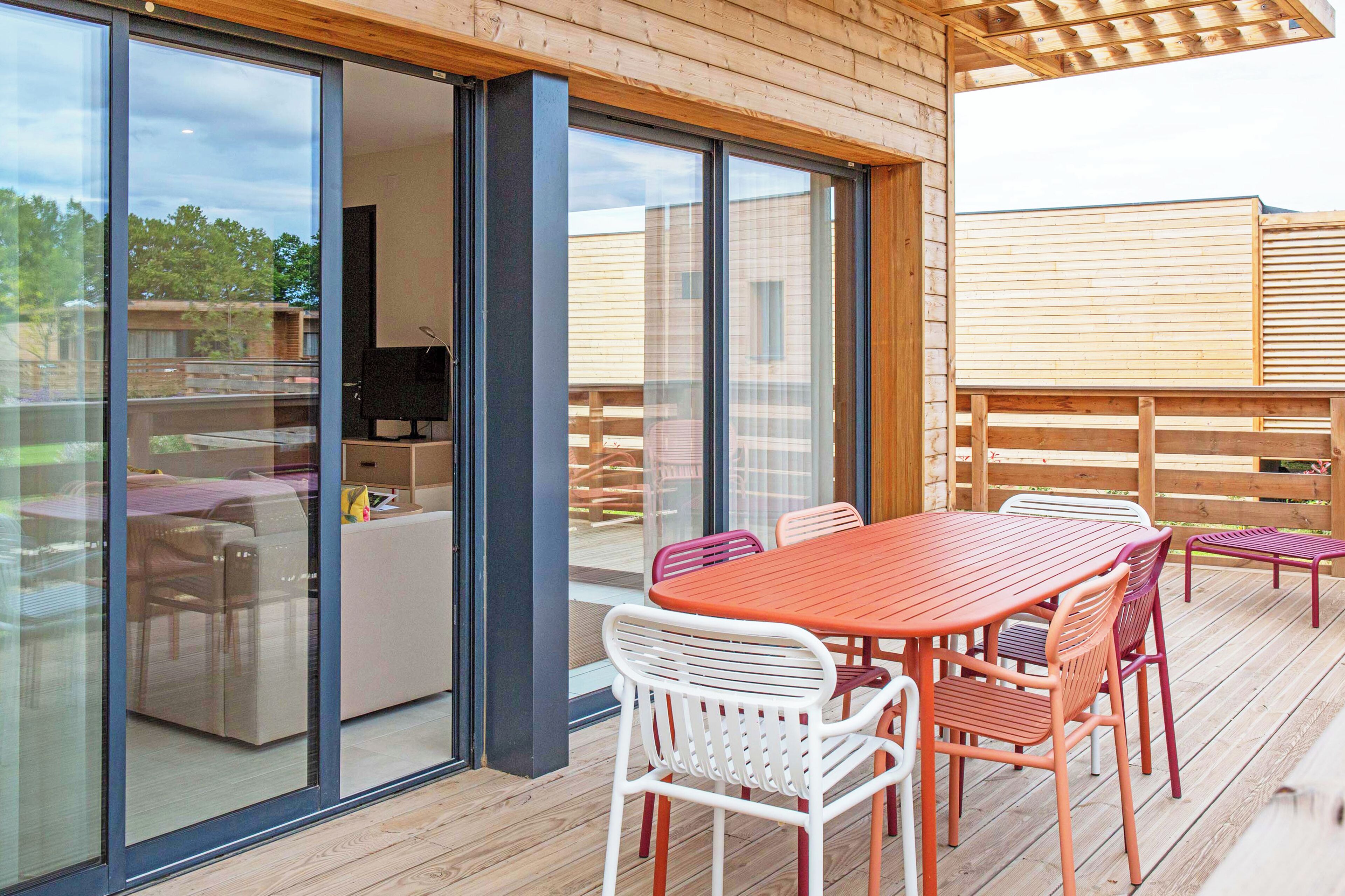 Eine Holzdielen-Terrasse mit Tisch und Stühlen, eine Glastür zu einem Zimmer und Blick auf die Umgebung.