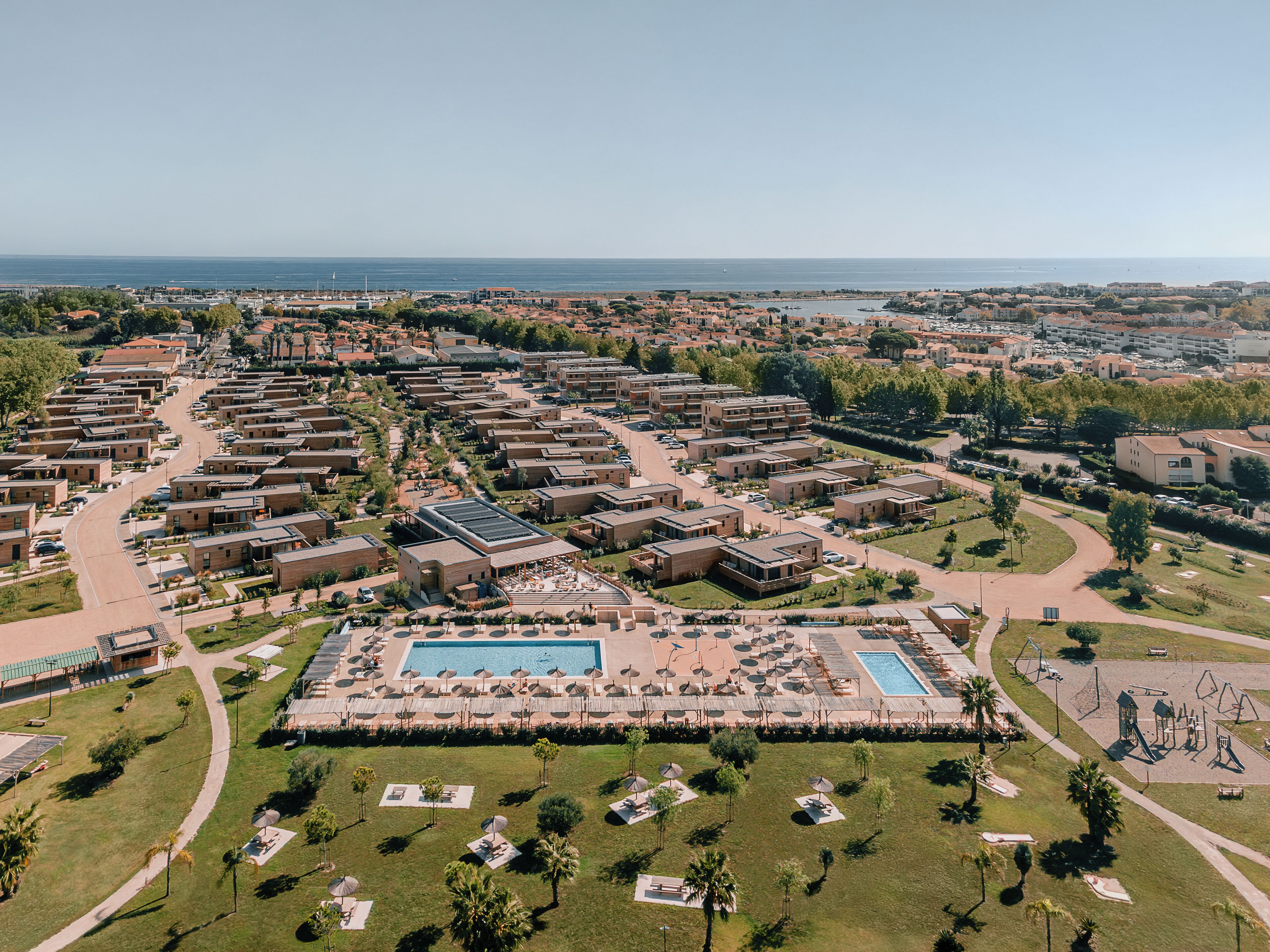 Vue aérienne d’un complexe hôtelier comprenant plusieurs bâtiments, un espace central avec une piscine et des chaises longues, et une vue sur la mer.