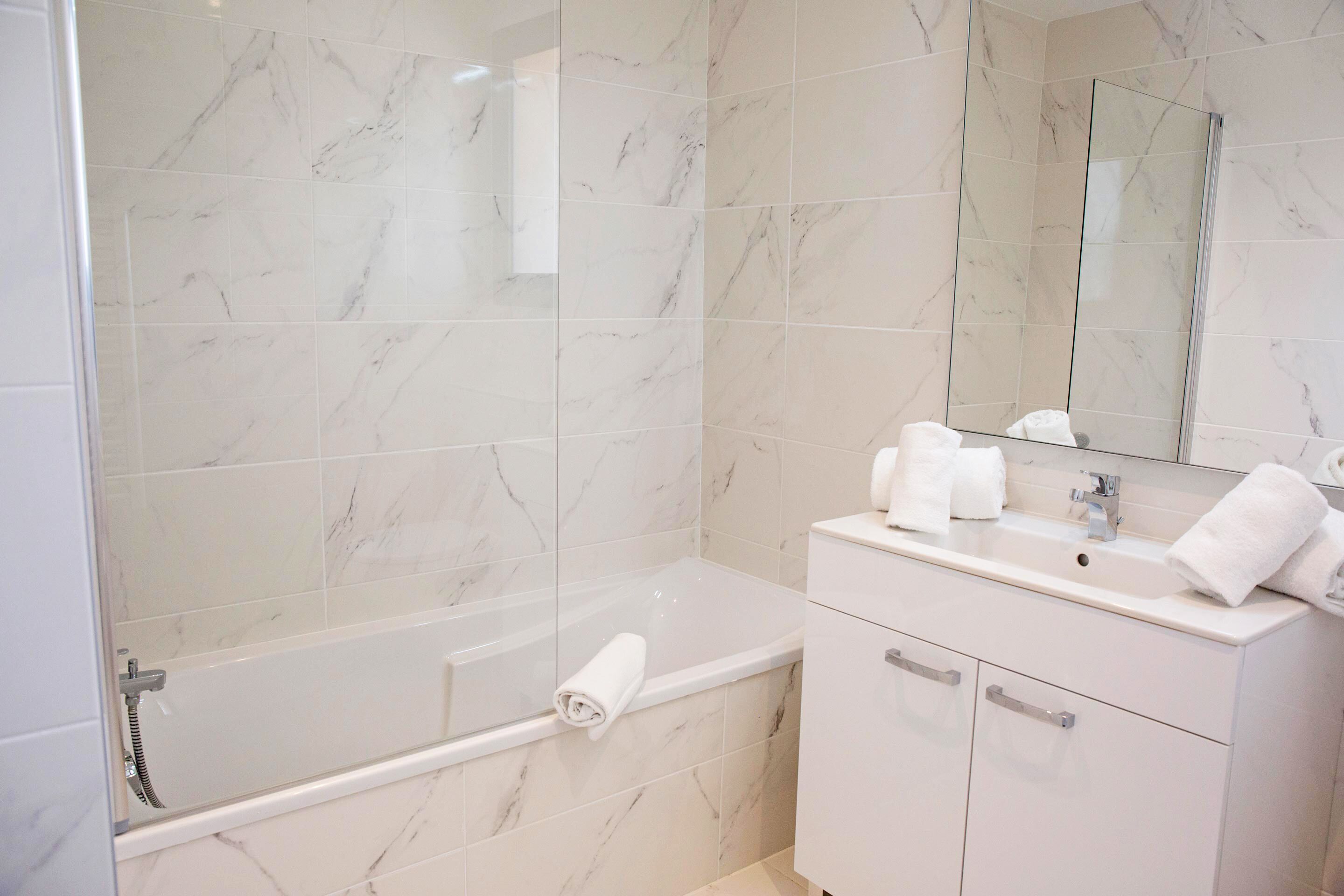 A modern bathroom with a glass-enclosed shower, a white marble-tiled wall, a white vanity with a mirror, and a towel rack.