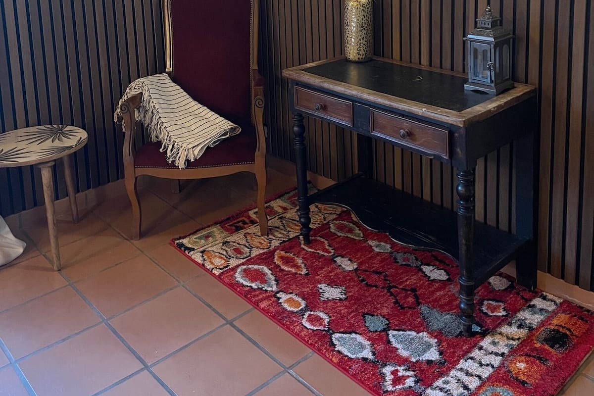 Una habitación con una silla de madera, una mesita pequeña, un escritorio, un tapete estampado y un espejo en la pared.