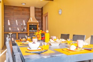 Un coin repas avec un four en briques, des murs jaunes et une longue table dressée pour le petit-déjeuner.