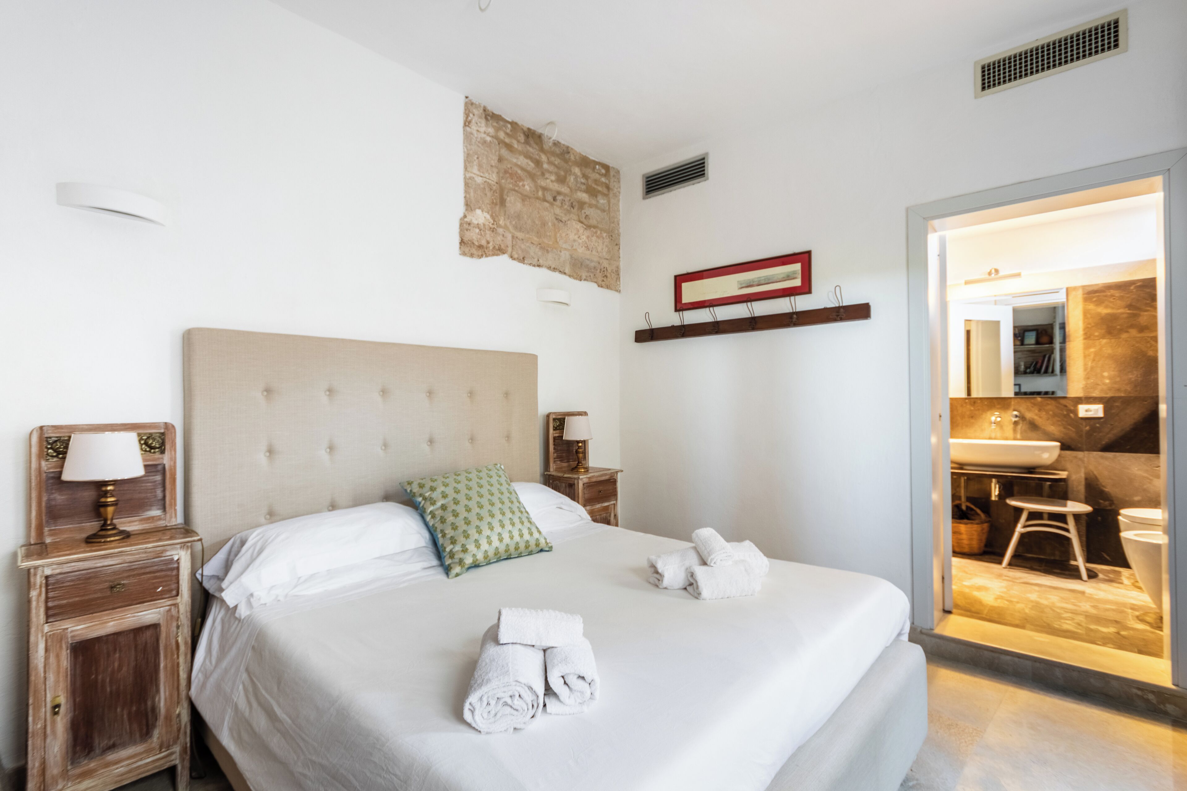 Une chambre d’hôtel avec un grand lit, une table de chevet avec une lampe, une étagère fixée au mur et une salle de bain visible à travers une porte ouverte.