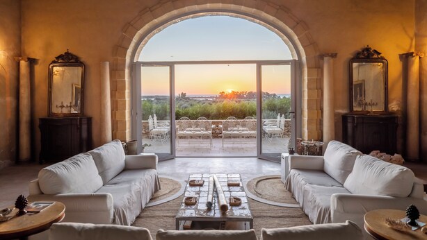 Um quarto espaçoso com uma grande janela arqueada com vista para um jardim e um pôr do sol. Há dois sofás brancos e uma mesa de centro no centro.