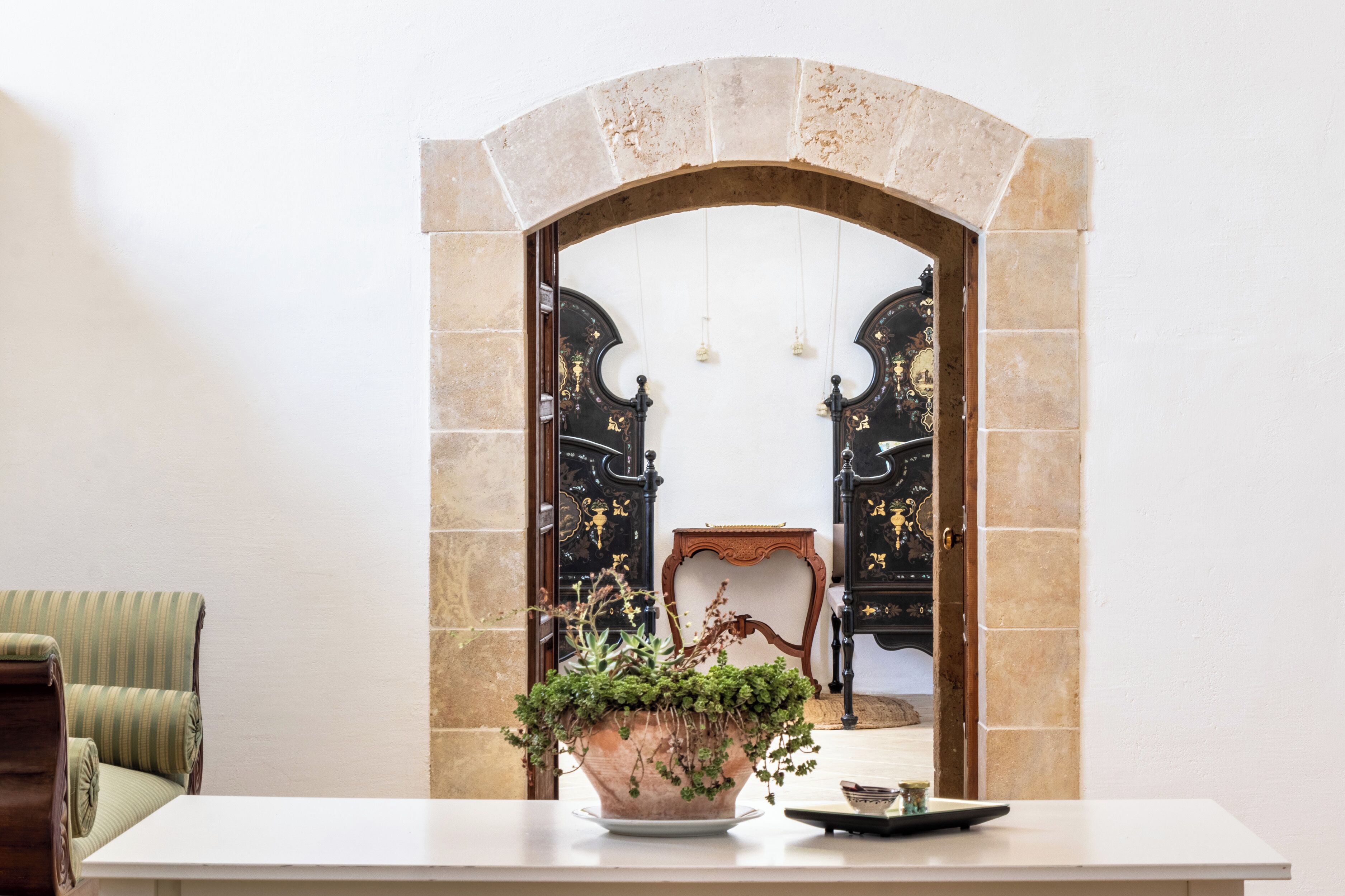 Um quarto com uma mesa branca, uma planta em vaso e vista através de uma porta arqueada para um quarto preto e dourado com uma cadeira e uma pequena mesa.