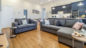 Ein modernes Wohnzimmer mit grauen Sofas, Holzboden und einer Küchenzeile mit dunklen Schränken und Pendelleuchten.