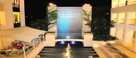 A modern hotel with a central water feature, outdoor seating, and a well-lit facade.