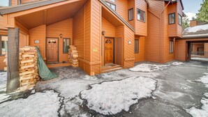 A two-story wooden house with a covered porch, a stack of firewood, and a snow-covered driveway. - Mammoth Lakes Condo: Walk to Village & Gondola! (Mammoth Lakes)