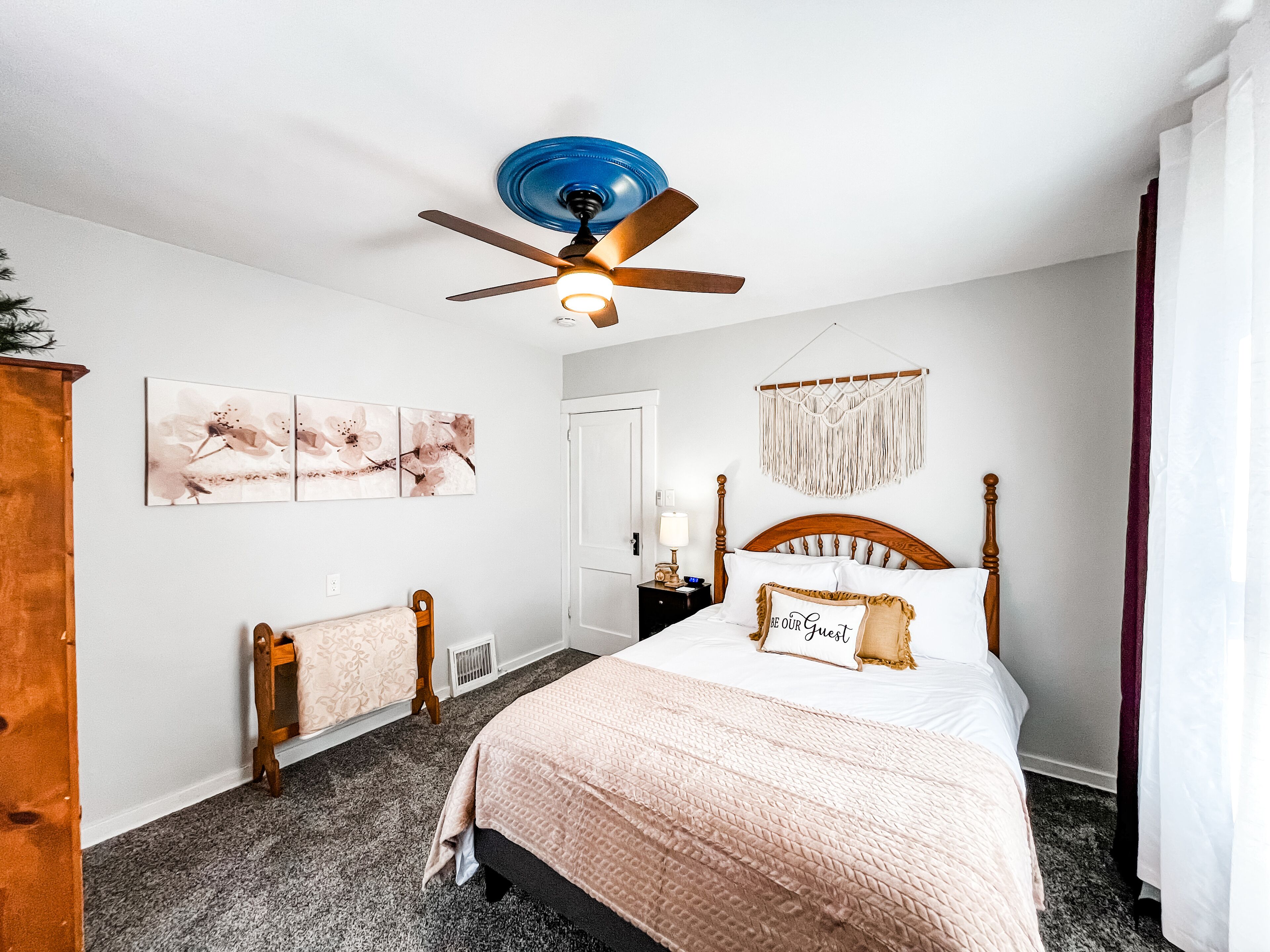 A bedroom with a bed, a ceiling fan, a nightstand, a wall-mounted macrame, and framed floral artwork.