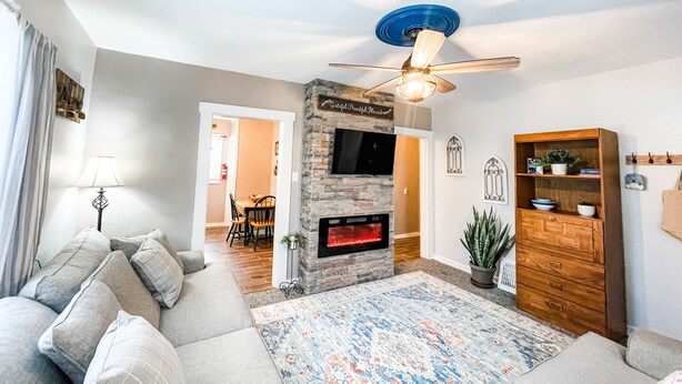 A living room with a stone fireplace, a ceiling fan, a wooden cabinet, and a dining area in the background.