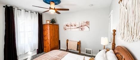 Ein Schlafzimmer mit einem Bett, einem Deckenventilator, einem Kleiderschrank, einem Fenster mit Vorhängen und Wandbildern.