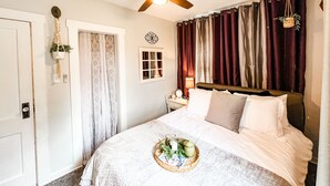 A bedroom with a bed, a ceiling fan, a window, curtains, a bedside table with a lamp, and a tray with items on the bed.