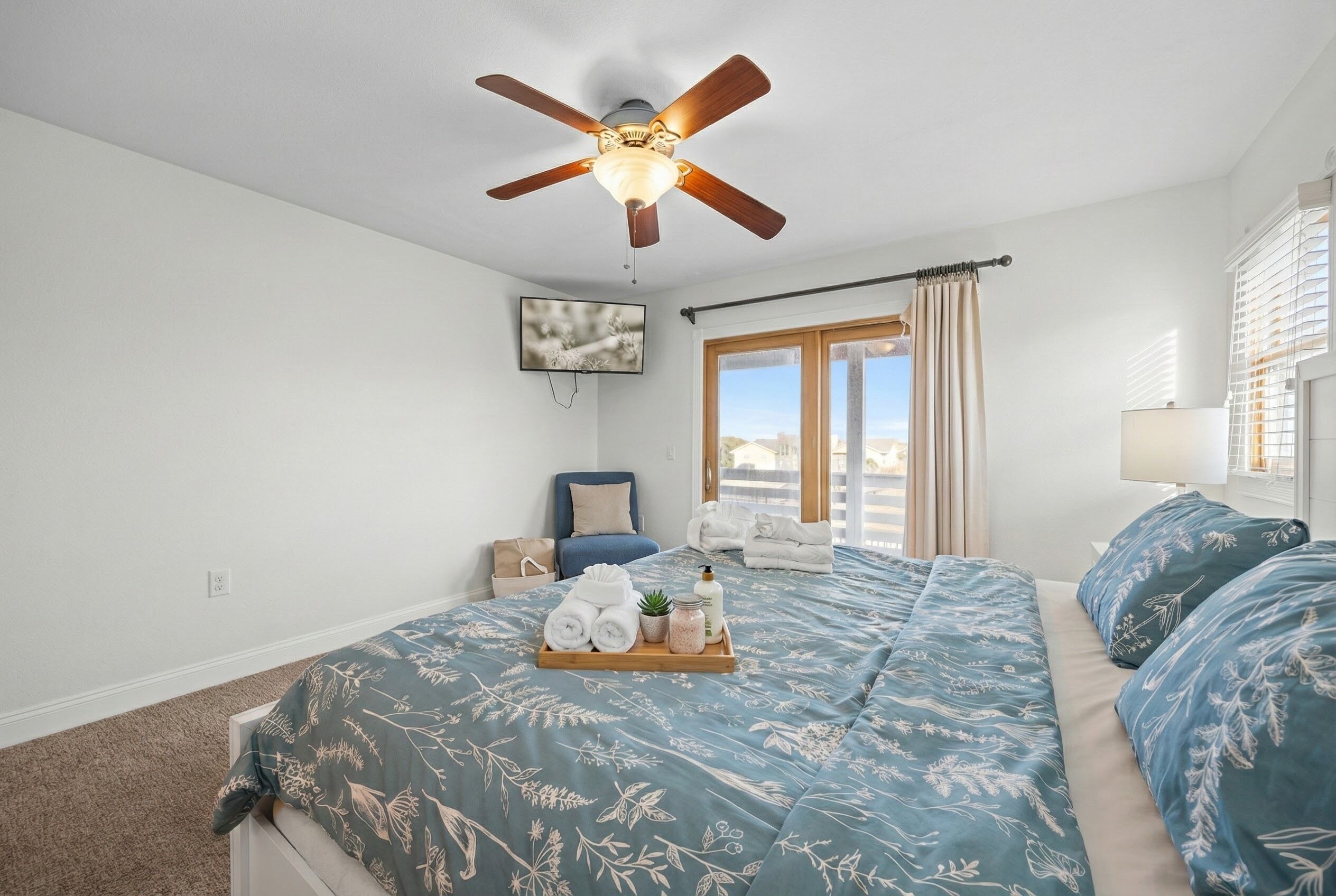 Una camera da letto con una ventola a soffitto, un letto con biancheria da letto a motivi floreali, un comodino con una lampada e vista all'esterno attraverso una finestra.