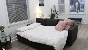 Une chambre à coucher moderne avec un grand lit, une table de chevet, une lampe et une vue sur la rue par la fenêtre.