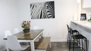 Un coin repas avec une table en bois, des chaises blanches, un banc et une œuvre d’art à motifs zébrés accrochée au mur.