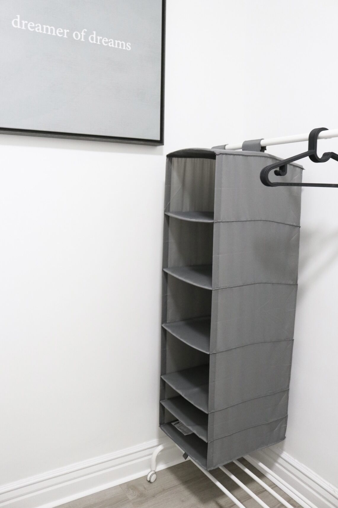 Un módulo de almacenamiento gris con varias repisas, fijado a una pared con un zócalo blanco y un cuadro enmarcado encima.