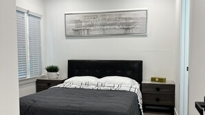 Une chambre à coucher comprenant un lit, une table de chevet, un tableau accroché au mur, une fenêtre avec des stores et une porte.