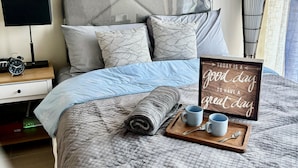 Una cama bien hecha con una toalla enrollada, una mesita de noche con lámpara y reloj, y un letrero motivacional en la cama.