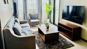 Una sala de estar moderna con un sofá, un sillón, una mesa de centro, televisor y ventanas grandes con cortinas.