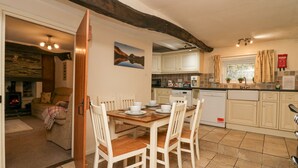 A cozy kitchen with a dining area, wooden chairs, a table set for four, and a view into a living room with a fireplace. - Taylor's Cottage (Keswick)