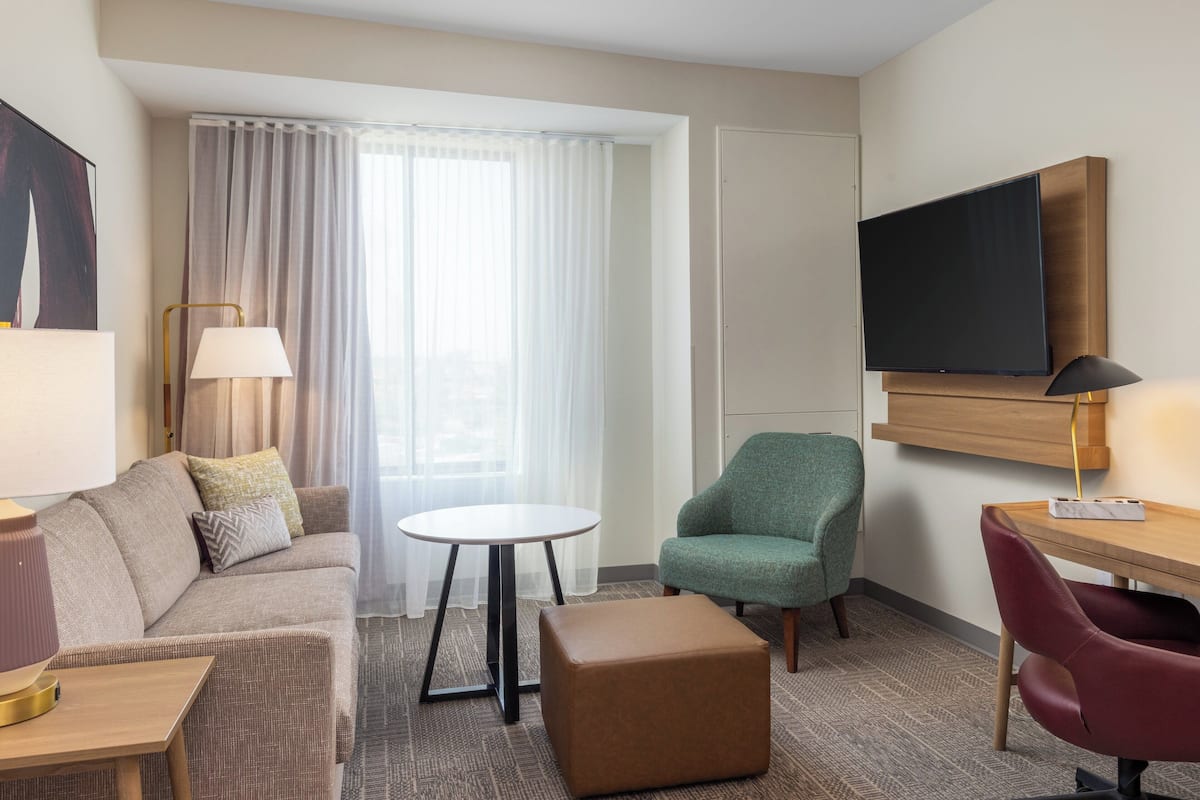 Habitación de hotel con sofá, una pequeña mesa redonda, un sillón verde, un escritorio de madera, un televisor en la pared y un amplio ventanal con cortinas.