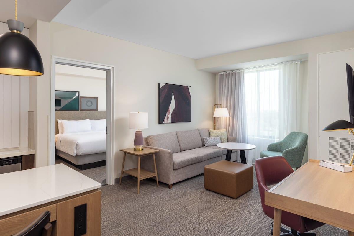 Una habitación de hotel con cama, sofá, mesa de comedor, silla, lámpara, un cuadro en la pared y ventana con cortinas.