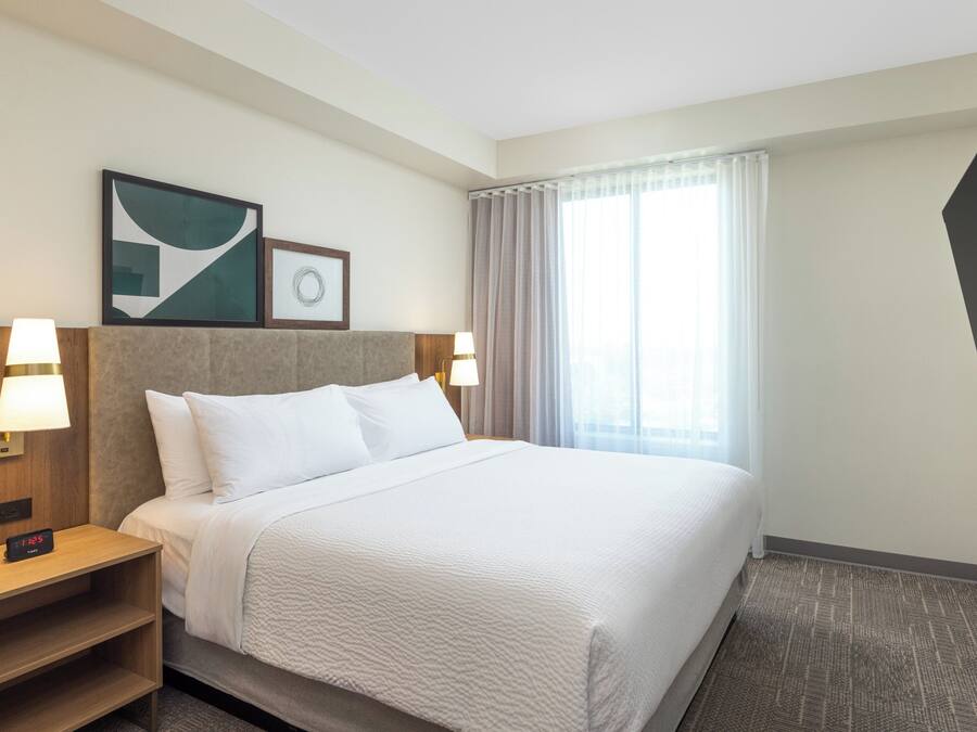 Una habitación de hotel con una cama grande, dos mesitas de noche con lámparas, un televisor montado en la pared y una ventana con cortinas.