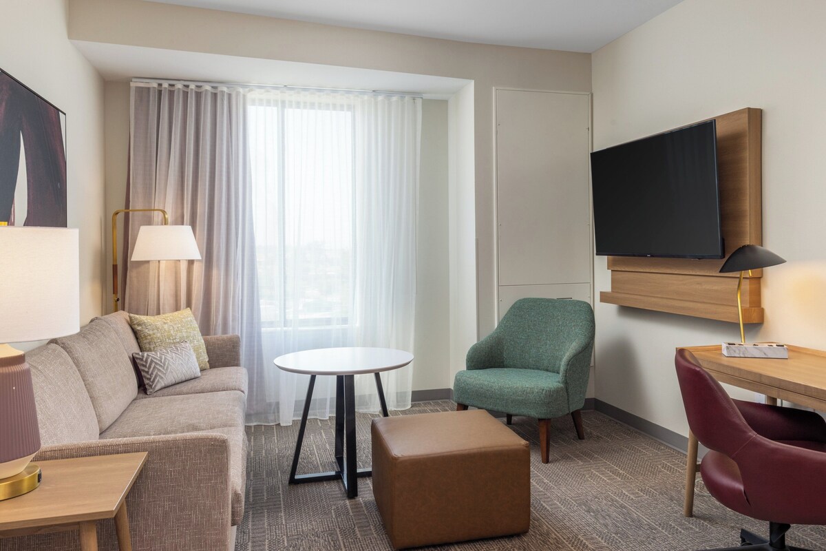 Habitación de hotel con sofá, una pequeña mesa redonda, un sillón verde, un escritorio de madera, un televisor en la pared y un amplio ventanal con cortinas.