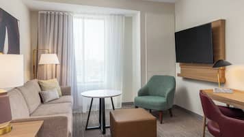 A hotel room with a sofa, a small round table, a green armchair, a wooden desk, a television mounted on the wall, and a large window with curtains.