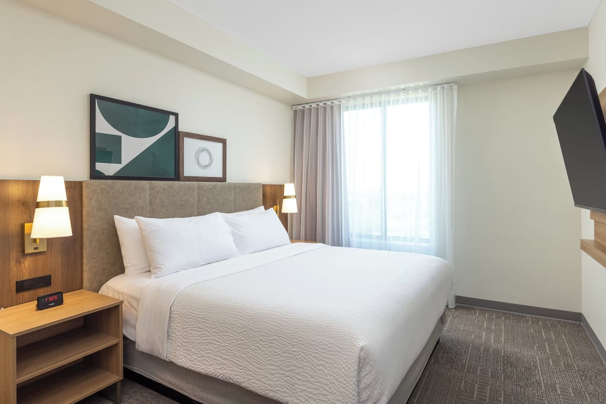 Una habitación de hotel con una cama grande, dos mesitas de noche con lámparas, un televisor montado en la pared y una ventana con cortinas.