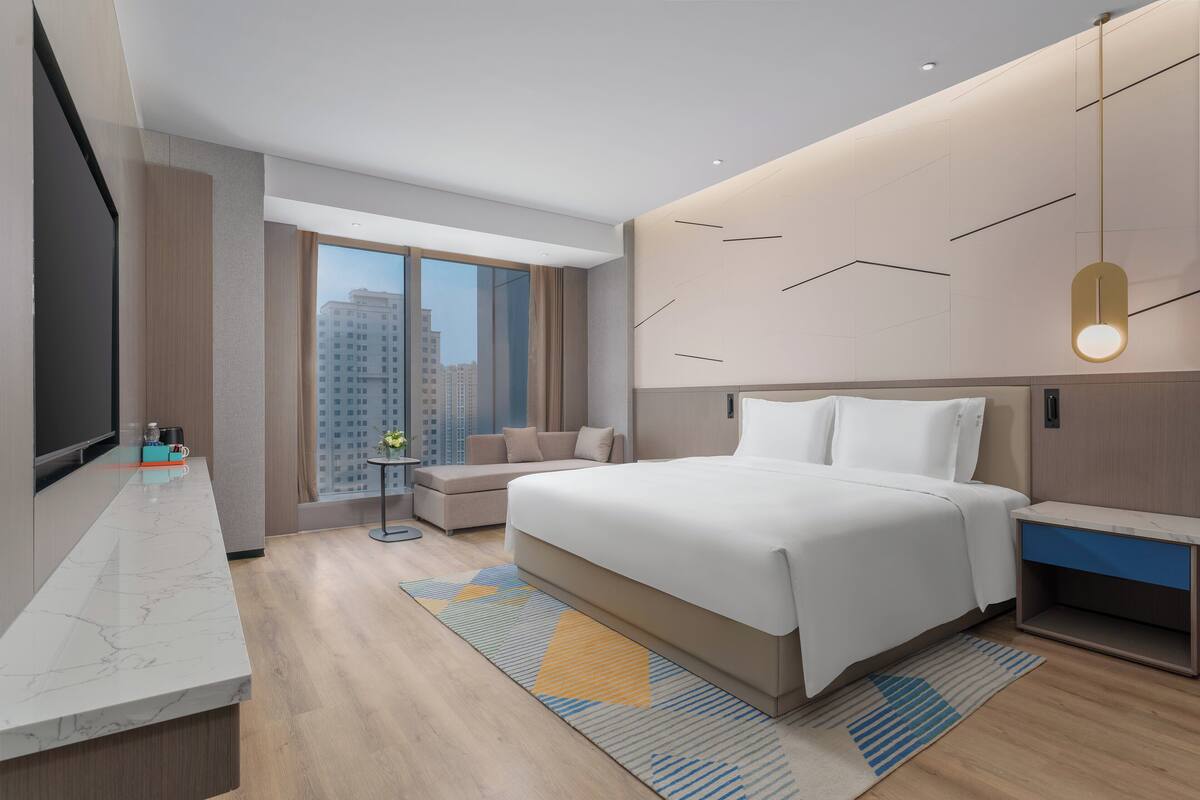 A modern hotel room with a large bed, a TV, a sofa, and a city view.