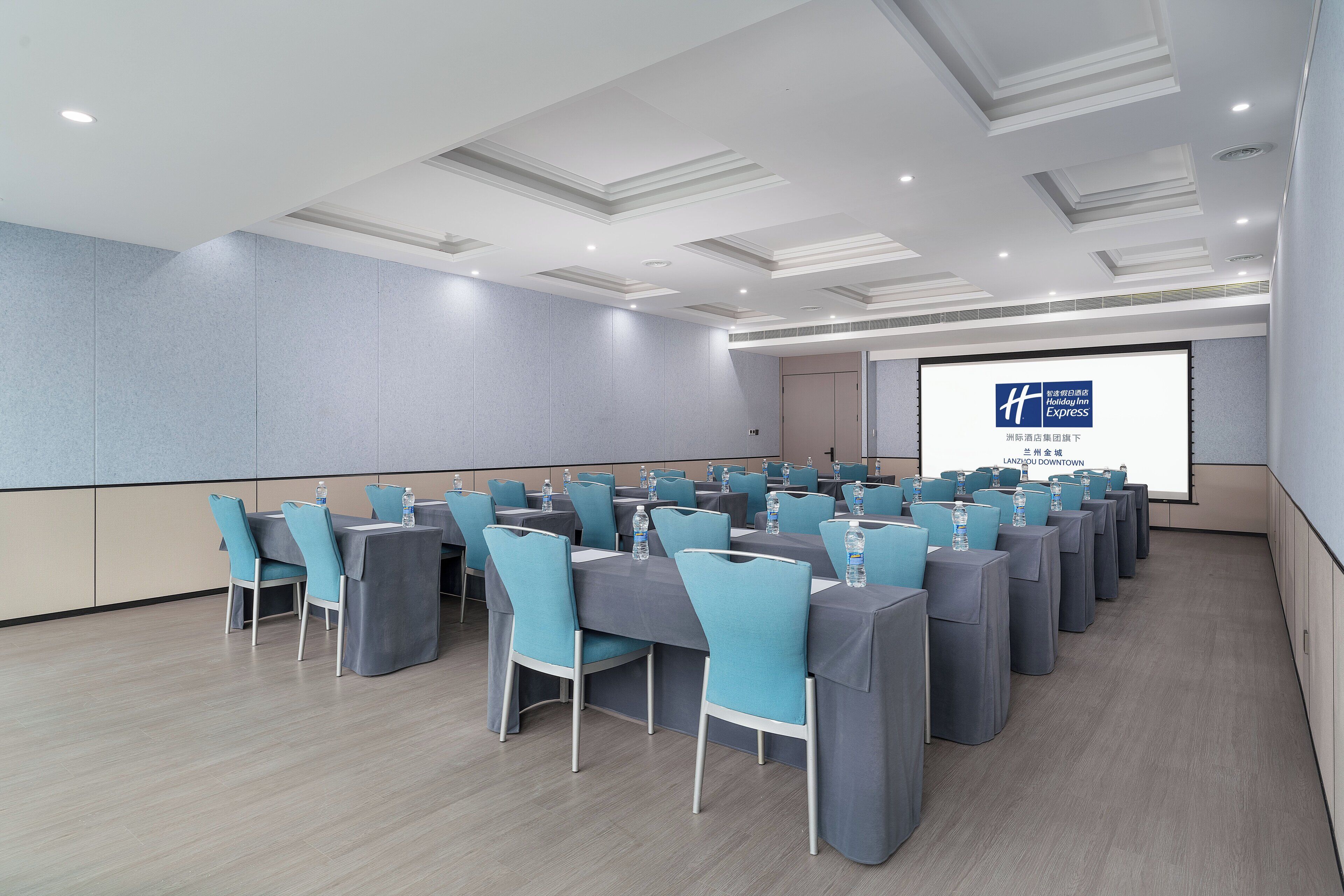 Un salón de conferencias con filas de mesas y sillas, una pantalla de proyección y un suelo de color claro.