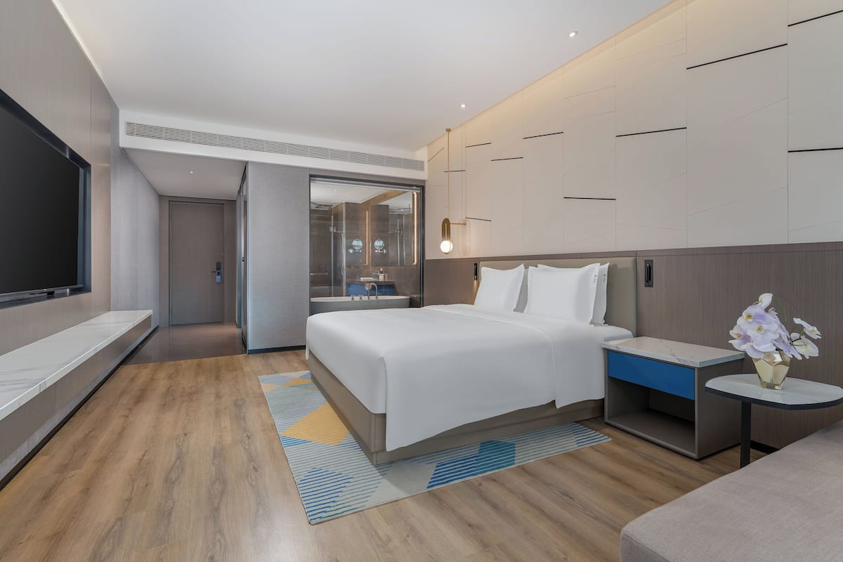 Habitación de hotel moderna con una cama grande, una mesita de noche con un florero y un televisor de pantalla plana, y vista al baño.