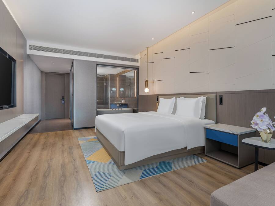 Een moderne hotelkamer met een groot bed, een nachtkastje met een bloevas, een flatscreen televisie en uitzicht op de badkamer.