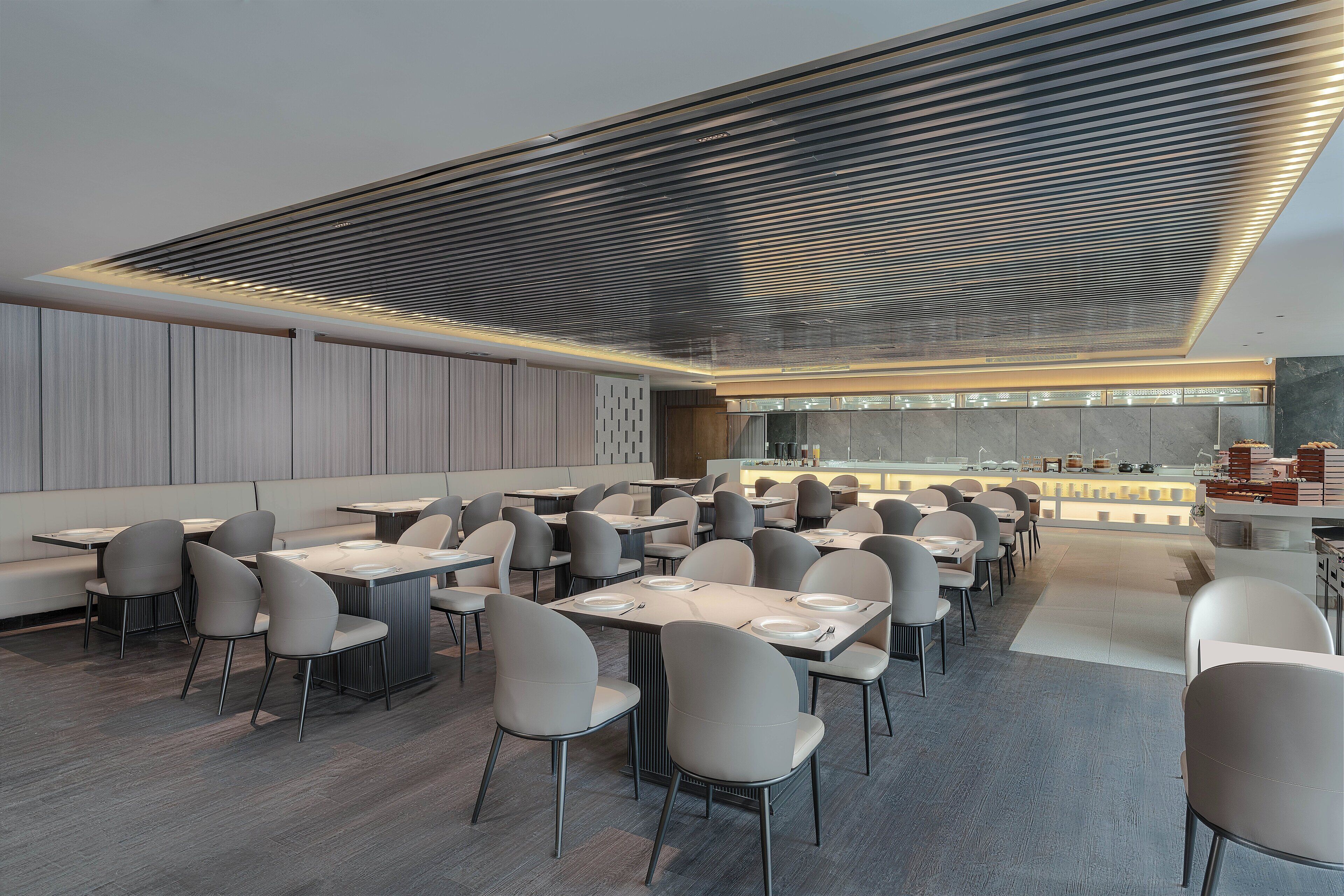 A modern restaurant with a long dining area, high ceiling with a patterned design, and a kitchen visible in the background.